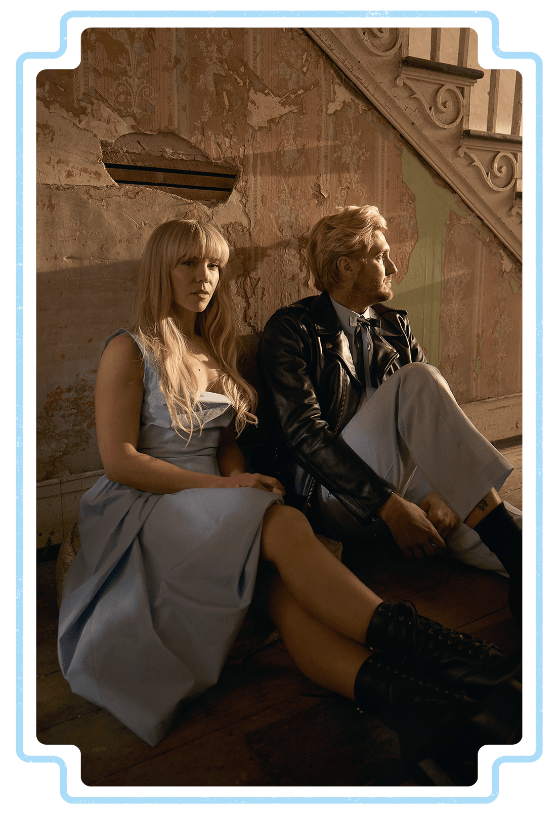 A man and woman sitting on the floor against a weathered wall with peeling paint in an old building. The woman has long blonde hair and wears a light gray dress and black lace-up boots. The man has blond hair, wears a black leather jacket, cream-colored pants, and black shoes, with his arms crossed and looking to the right.